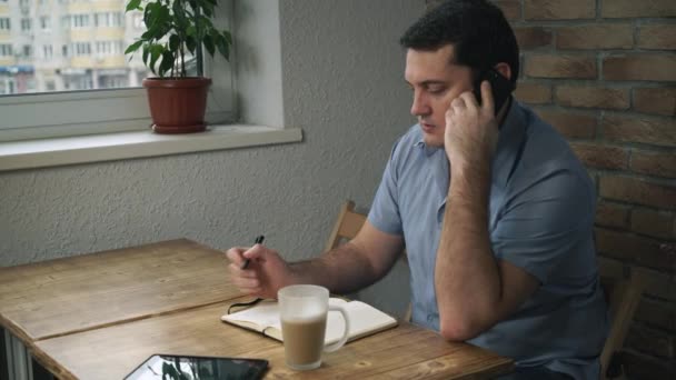 Gerente sentado en el café, hablando por teléfono, tomando notas en un bloc de notas . — Vídeo de stock