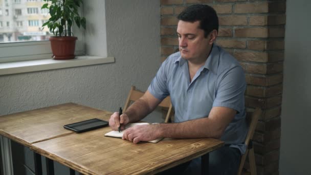 Hombre adulto sentado en un café con un diario y una tableta, tomando notas en un cuaderno . — Vídeo de stock