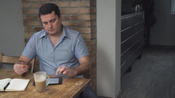 El hombre de mediana edad paga facturas a través de Internet en manos de la tarjeta de crédito. En la mesa hay una taza de café . — Vídeo de stock