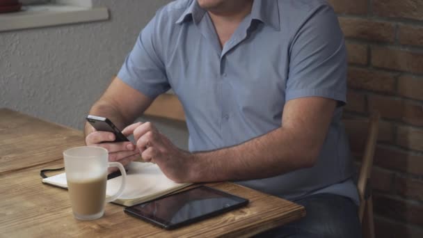 En man sitter vid ett skrivbord med en telefon i handen, ligger Tablet PC och bärbara. — Stockvideo