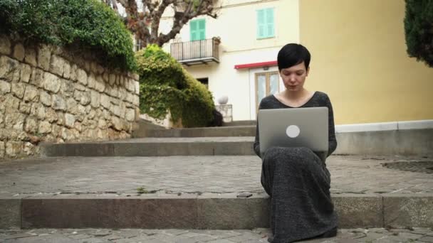 Hermosa mujer sentada en las escaleras usando la computadora frente a la casa de color . — Vídeo de stock