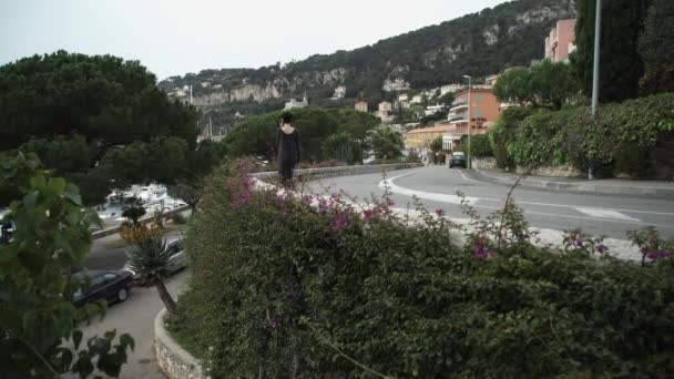 Mujer caminando por la carretera en la ciudad mediterránea a lo largo de verdes arbustos y flores . — Vídeo de stock