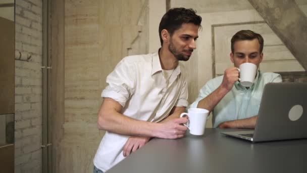 Dois jovens homens profissionais bebendo café olham para o computador e conversam — Vídeo de Stock