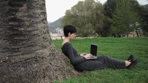 Vuxen kvinna som arbetar på datorn i parken under träd. — Stockvideo