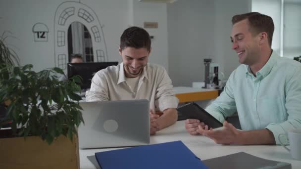 Due lavoratori sul posto di lavoro utilizzando un dispositivo moderno e chiacchierando di notizie divertenti . — Video Stock