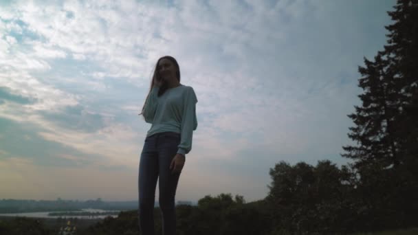 Das Mädchen mit den langen Haaren steht auf dem Hügel und telefoniert vor dem Hintergrund des Abendhimmels mit Wolken. — Stockvideo