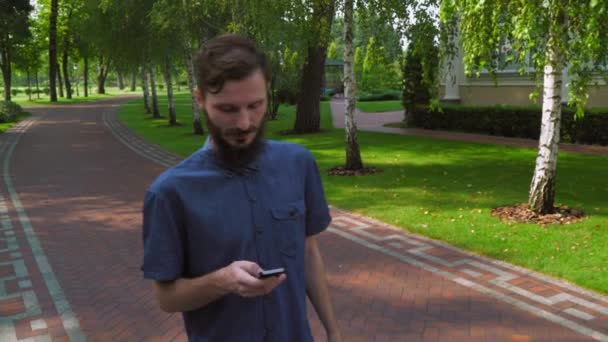 Un joven va por un camino en un parque cerca del edificio y recibe un mensaje por teléfono. . — Vídeos de Stock