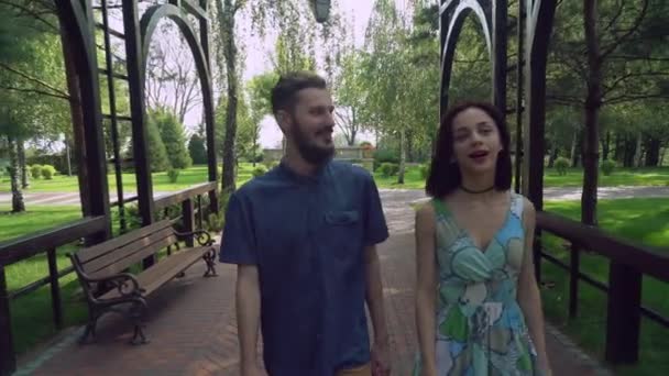 Pareja joven caminando por un sendero en el parque bajo el arco, charlando y sonriendo . — Vídeo de stock