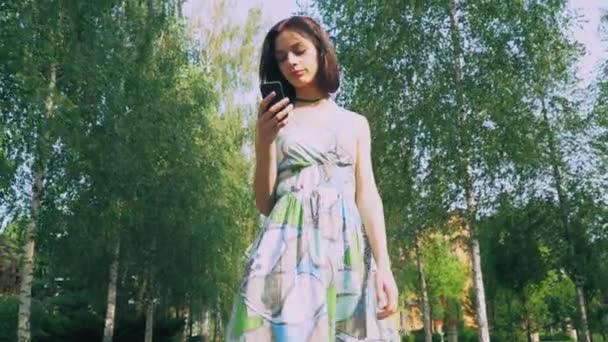 Chica joven en un vestido brillante está en el parque, leyendo un mensaje en su teléfono y sonríe . — Vídeo de stock