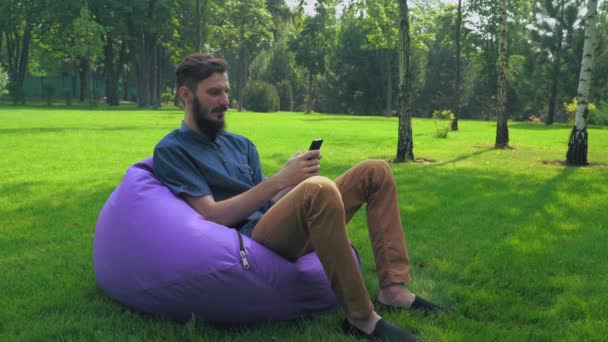 Ένας νεαρός άνδρας με ένα μπλε πουκάμισο κάθεται σε μια καρέκλα στο πάρκο και να αποκτήσουν ένα μήνυμα στο τηλέφωνο. — Αρχείο Βίντεο