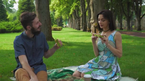 Jovem casal no parque de piquenique de verão para comer sanduíches, curtindo o sabor e sorrindo . — Vídeo de Stock