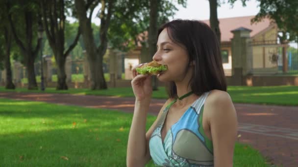 Chica sentada en el suelo en el parque y comiendo un sándwich . — Vídeos de Stock