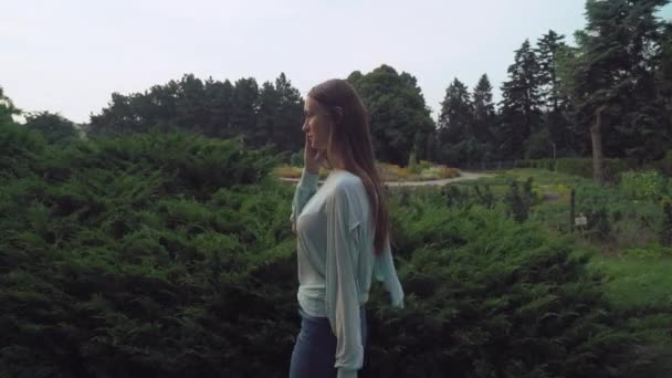若い女性が公園で携帯電話で話している茂みに沿って行く. — ストック動画