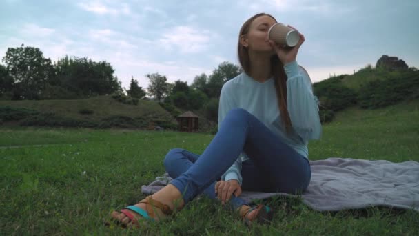 Uma jovem sentada no gramado do parque e bebe um copo de café . — Vídeo de Stock