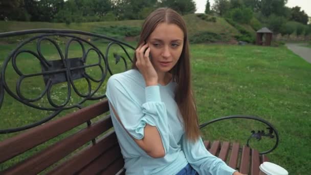 Uma jovem senta-se em um banco do parque, segurando uma bebida e falando ao telefone, sorri . — Vídeo de Stock