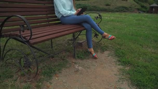 年轻的女孩坐在公园长凳和刷卡页面在 tablet 上. — 图库视频影像