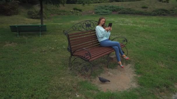 Chica joven con el pelo largo sentado en un banco del parque y sostiene la tableta, sonriendo . — Vídeos de Stock