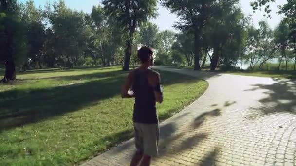 Un hombre joven con auriculares y ropa deportiva corre un día de verano, árboles verdes . — Vídeos de Stock