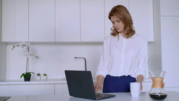 Businesswoman works in the kitchen with a laptop and drinks coffee Stock Picture