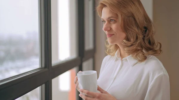 Frau steht mit einer Tasse Tee am Fenster lizenzfreie Stockbilder