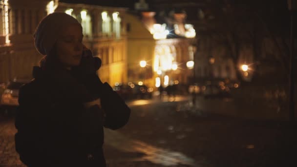 Жінка на вулиці вечора п'є каву і розмовляє по телефону — стокове відео