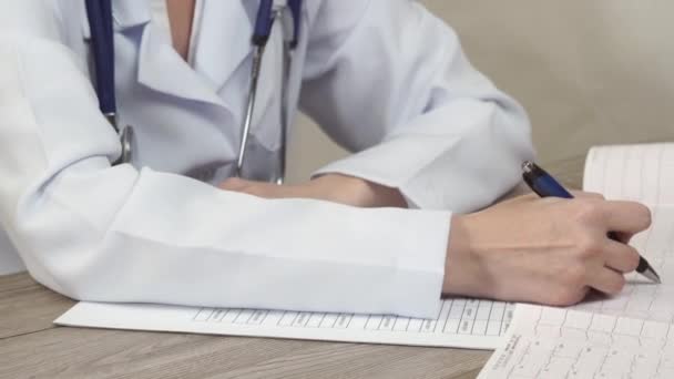 El médico mira el cardiograma y toma notas en un documento . — Vídeos de Stock
