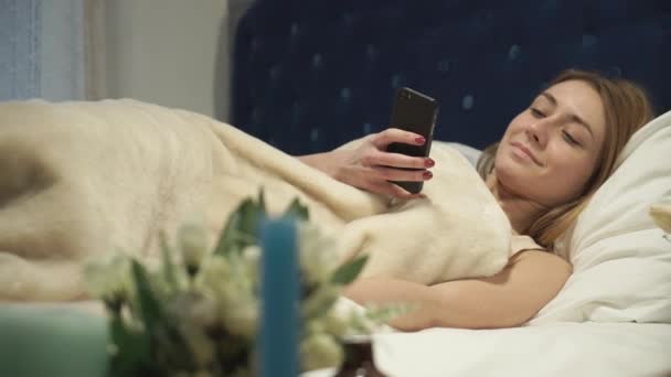 Ξανθιά ξαπλωμένη στο κρεβάτι, πληκτρολογώντας ένα μήνυμα στο τηλέφωνό σας — Αρχείο Βίντεο