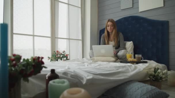 A menina em uma cama que trabalha com um laptop e café da manhã — Vídeo de Stock