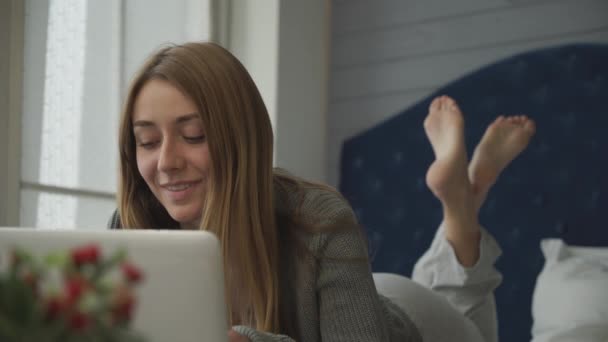 Chica sonriente acostada en la cama con un portátil — Vídeo de stock