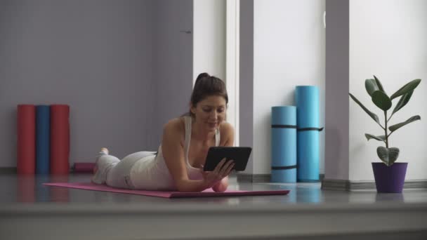 Femme couchée sur un tapis de yoga et regardant la tablette — Video