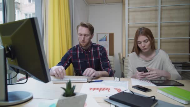 Man geconcentreerd werk op de computer, de vrouw op zoek naar slimme telefoon. — Stockvideo