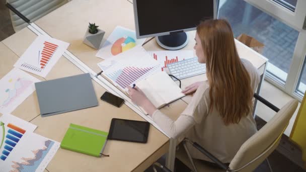 En kvinna som sitter på arbetsplatsen, att göra anteckningar, noggrant studera diagrammen. — Stockvideo
