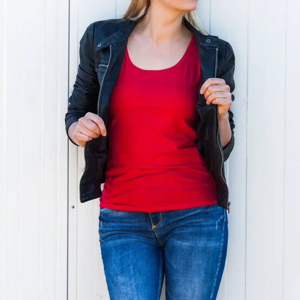 シティTシャツの白い金属ドアに対する赤Tシャツと黒の革のジャケットの下のボディ女性がモックアップ — ストック写真