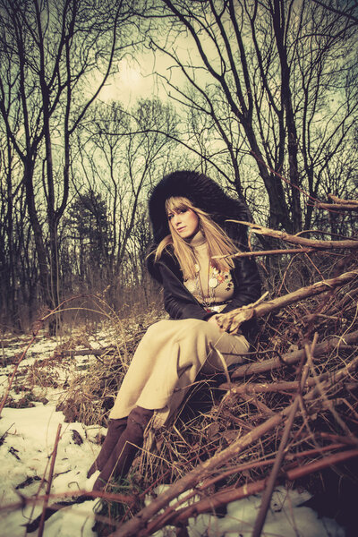 Blonde woman in coat with hood and long wool dress sit on branches in the forest, cold winter day, retro colors