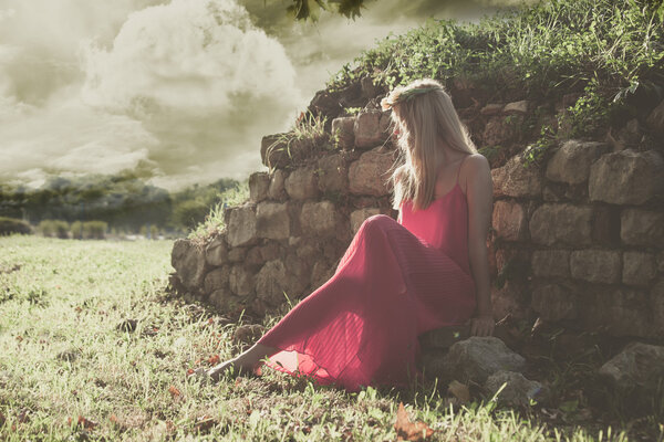 Fairy like young woman in red dress and wreath of flowers in hair sit by old wall in nature, full body shot, side view