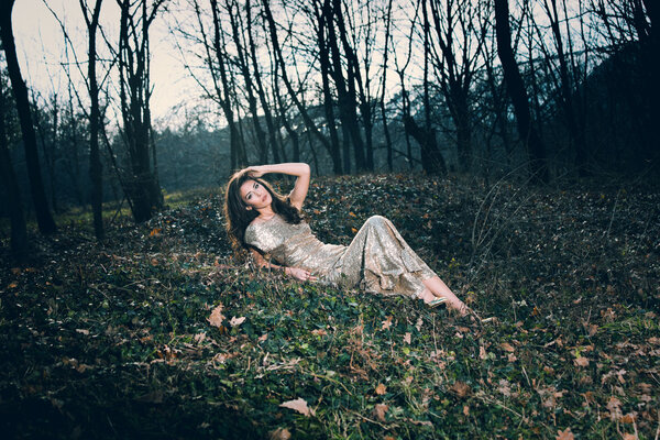 Beautiful young woman in elegant glittering dress in wood lie on leaves full body shot