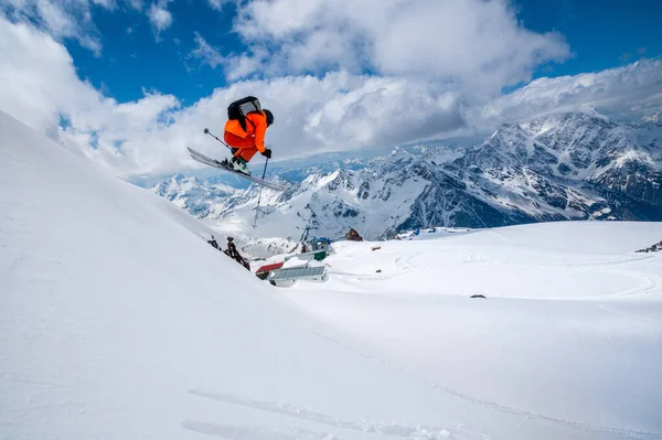 Επαγγελματίας αθλητής σκιέρ freerider σε ένα πορτοκαλί κοστούμι με ένα σακίδιο πετά στον αέρα μετά το άλμα στις υστερίες στο φόντο του μπλε χιόνι και τα βουνά — Φωτογραφία Αρχείου