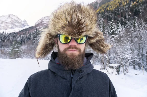 Retrato de um atraente homem sorridente barbudo em óculos de sol e um chapéu de pele contra o fundo de uma floresta nevada — Fotografia de Stock
