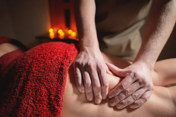 Close-up mãos masculinas de um massagista fazendo uma massagem na coxa para uma cliente mulher em um quarto escuro de um salão de spa em um fundo de velas — Fotografia de Stock