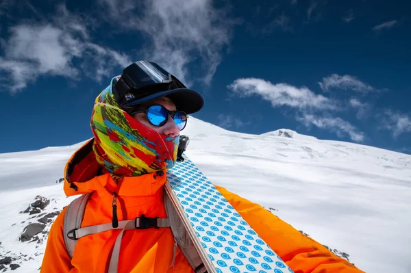 Güneş gözlüklü sert bir kayakçı portresi ve yüzünde kar maskesi olan bir şapka. Kayaklarını omzunda tutuyor ve Elbrus Dağı 'na bakıyor. — Stok fotoğraf