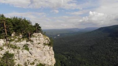 Uzun saçlı, sıska, siyah tişörtlü ve şortlu bir adam uçurumun kenarında duruyor ve etrafındaki orman ve dağlara bakıyor. Hava görünümü