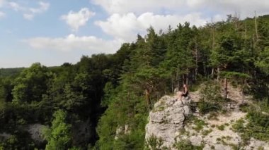 Uzun saçlı, siyah tişörtlü ve şortlu zayıf bir adam uçurumun kenarında oturur ve etrafındaki orman ve dağlara bakar. Hava görünümü