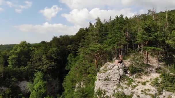 A long-haired, thin man in a black T-shirt and shorts sits on the edge of a cliff and looks out at the surrounding forest and mountains. Aerial view — Stock Video