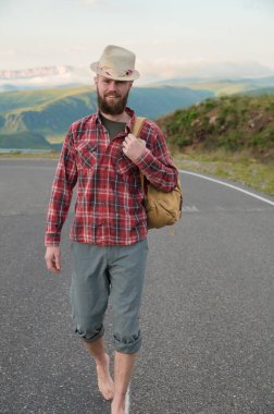 Sırt çantalı genç otostopçu erkek turist gün batımında dağların arka planına karşı asfalt yolda yürüyor.