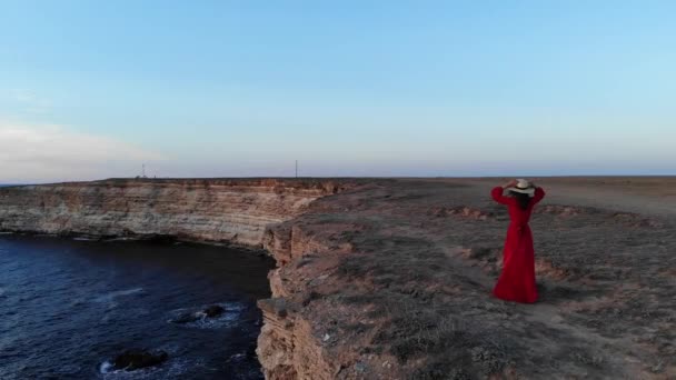 Aerial view of a young woman in a red dress and a straw hat walks along the edge front of a cliff by the sea alone at sunset — Stock Video