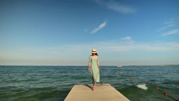 緑の夏のドレスを着た魅力的な白人の女の子と、晴れた日には海の海岸の木製の桟橋に沿ってわら帽子が歩いています。海で休みなさい。旅行ビデオ — ストック動画