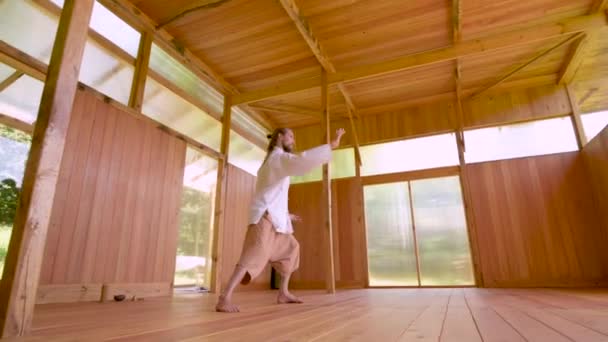 En långhårig vit man i lätt och lös klädsel övar qigong tai chi i ett övningsrum i trä på sommaren. Långsamma rörelser harmoni och lugn — Stockvideo