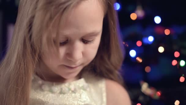 축제의 반짝 이는 드레스를 입은 한 어린 소녀가 반짝이는 신년 조명을 배경으로 아래를 보고 — 비디오