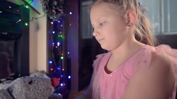 Pequeña chica gorda alegre y solitaria jugando con una rata de juguete suave en el fondo de una chimenea eléctrica y luces parpadeantes de año nuevo en la noche de Navidad — Vídeos de Stock