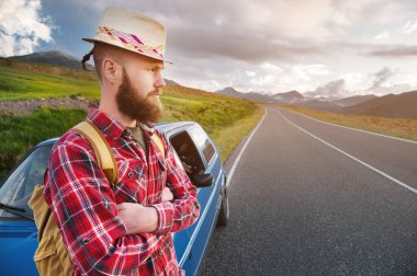 Çekici beyaz sakallı, şapkalı ve sırt çantalı ekose gömlekli adam güzel bir dağ manzaralı, kır yolunun kenarında arabasının yanında duruyor.